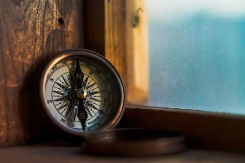 A vintage-style compass resting on a wooden windowsill, softly lit by natural light coming through a slightly frosted window.