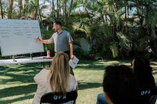 Jason Rowinski leads an outdoor session, standing beside a large whiteboard and writing as several participants sit facing him. He holds a paper in one hand, and the setting is surrounded by tropical plants under clear daylight.